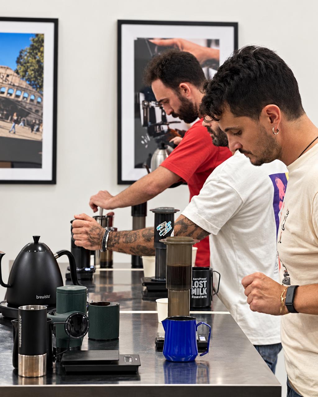Finalists brewing inverted AeroPress side by side at a steel bar with gooseneck kettles, hand grinders and scales