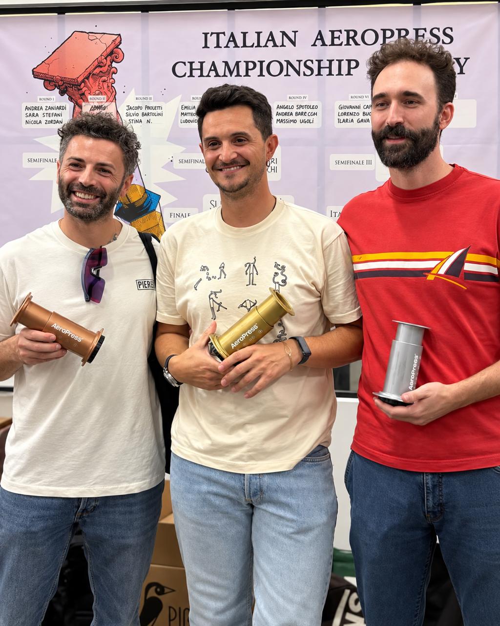 The three finalists of the 2026 Italian AeroPress Championship holding copper, gold and silver AeroPress trophies in front of the competition bracket