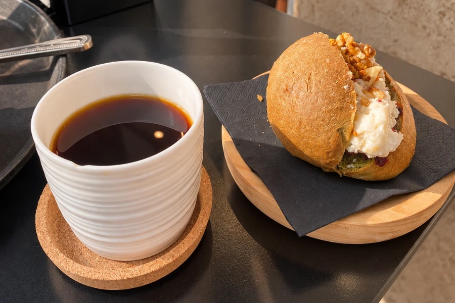 Honduras filter coffee in a white ceramic cup on a cork coaster, next to a spinach bauletto filled with fresh cheese, walnuts and cranberries