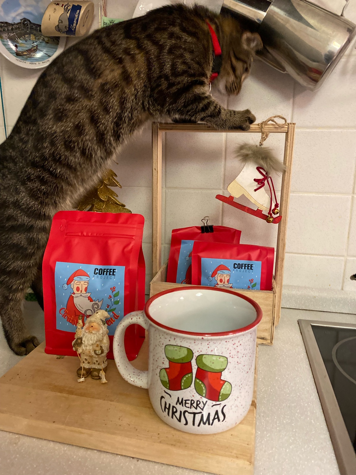 A tabby cat reaching over a wooden crate with an ice-skate ornament, above red Christmas coffee bags, a Santa figurine and a Merry Christmas mug on a kitchen counter