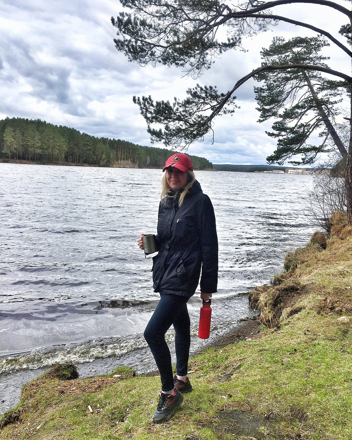 Sasha Pavlovich holding a metal camping mug of coffee on a lakeshore under a pine tree