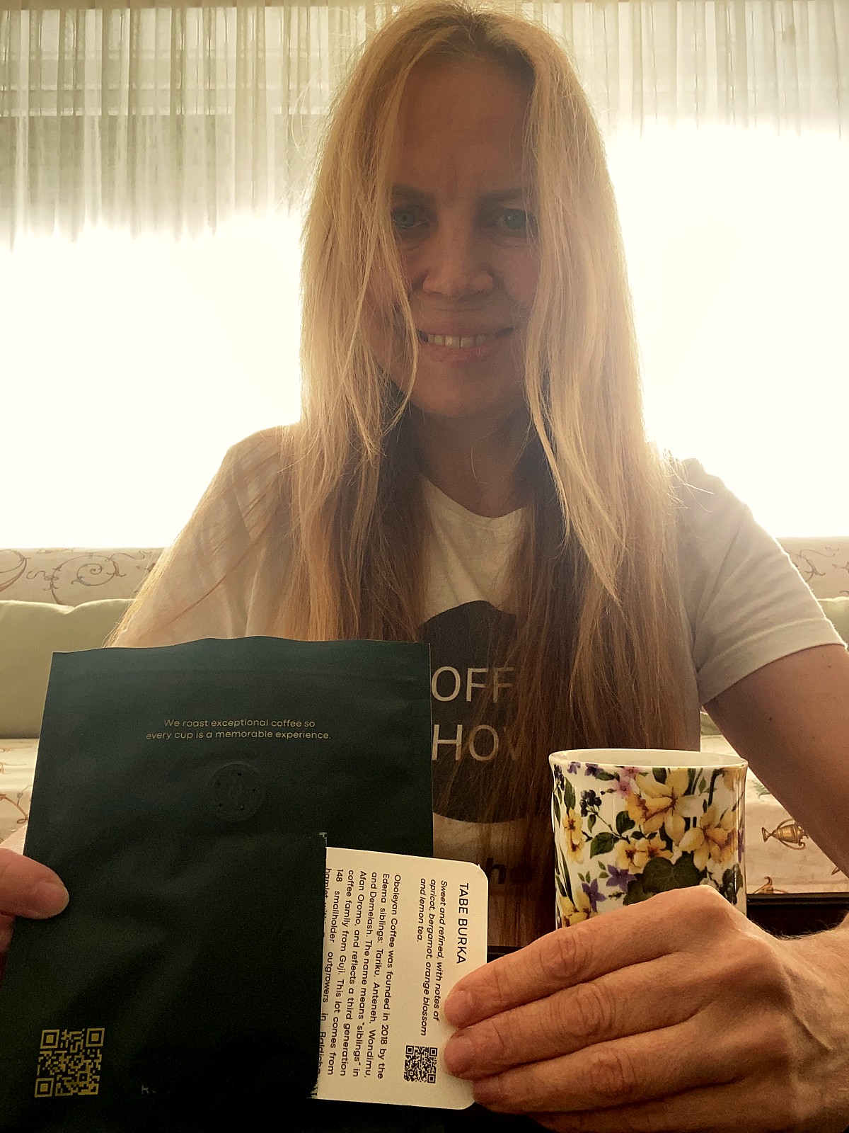 Sasha Pavlovich holding the back of the Rozali Coffee bag with the Tabe Burka lot card in its pocket, a floral mug beside her