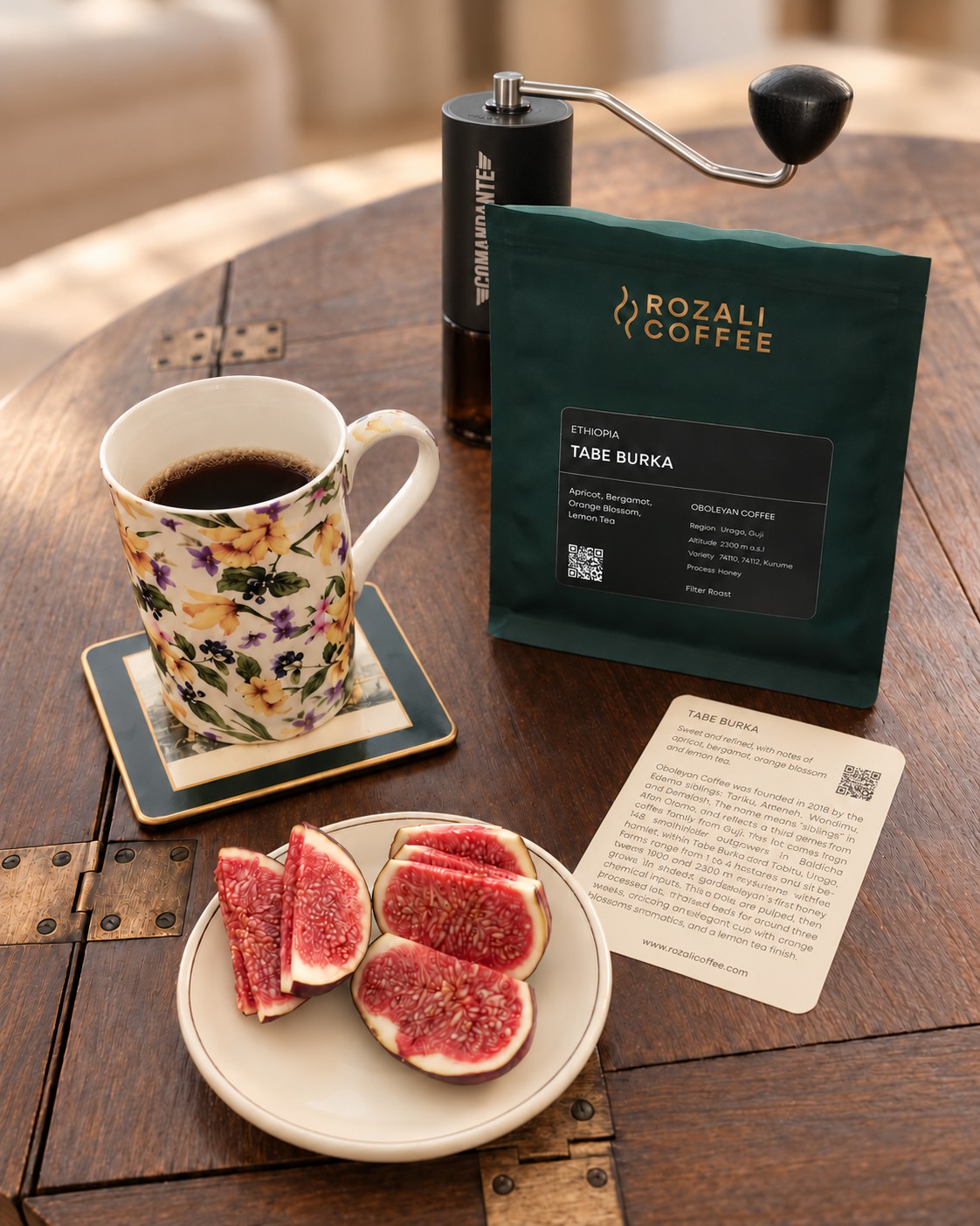 Mug of black coffee beside a Comandante hand grinder, a Rozali Coffee Tabe Burka bag, its lot card and sliced figs