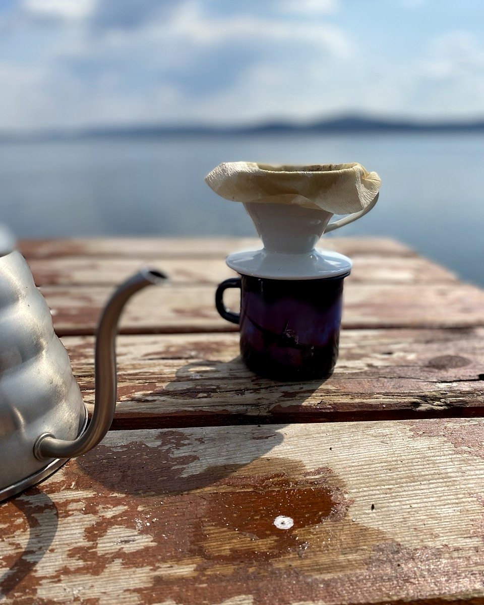 Ceramic V60 dripper on an enamel mug with an oversized Chemex paper filter folded down over its rim, a gooseneck kettle beside it and a lake behind
